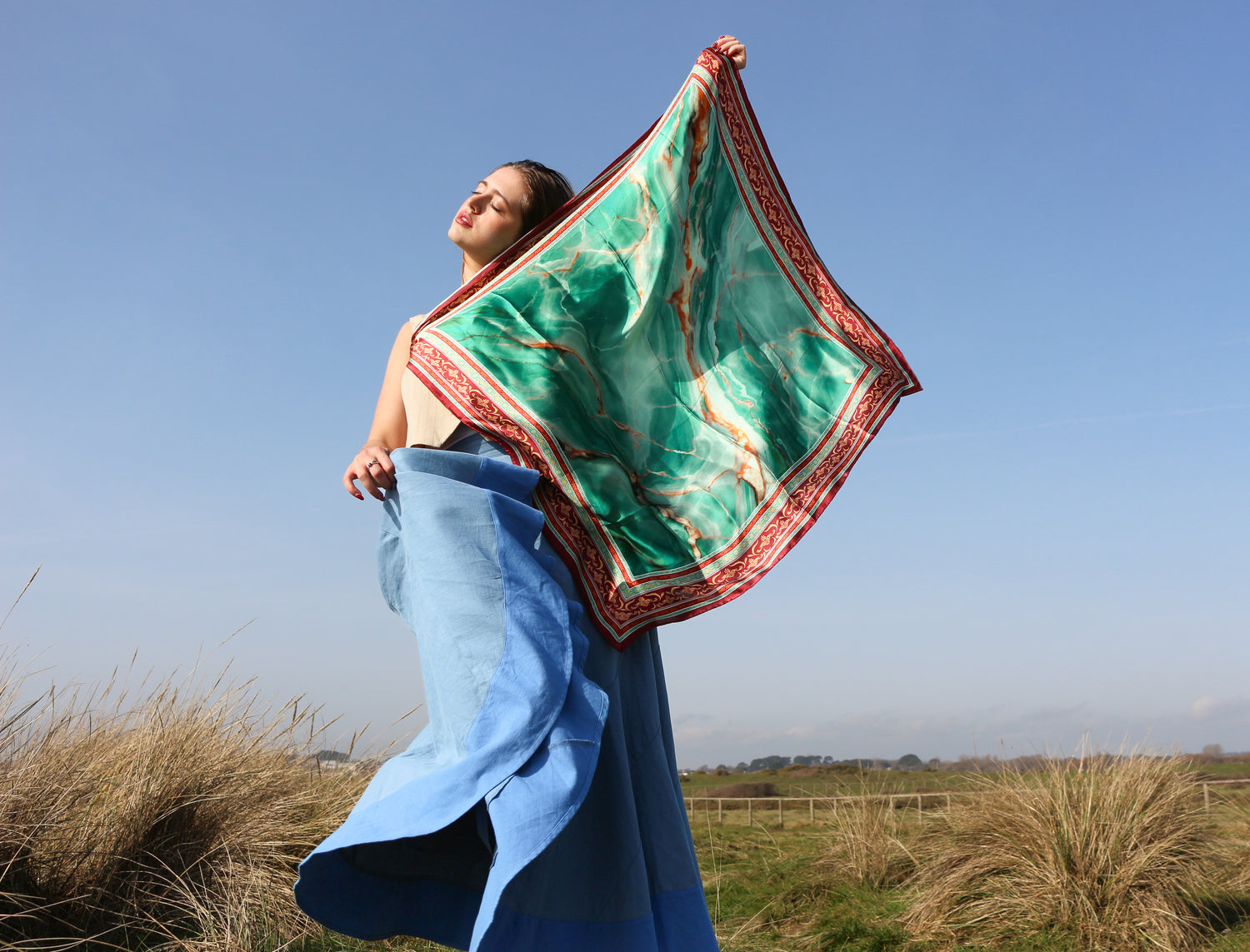 Woman holding a green and red patterned scarf in an open field with a clear blue sky.